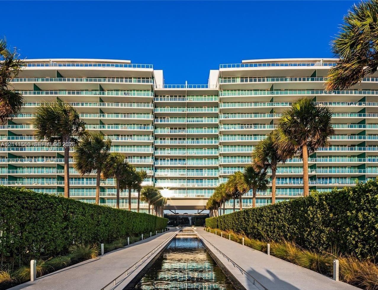 Multi-story residential building featuring expansive floor-to-ceiling glass balcony railings, a central reflecting pool, paved pedestrian walkways, and manicured landscaping