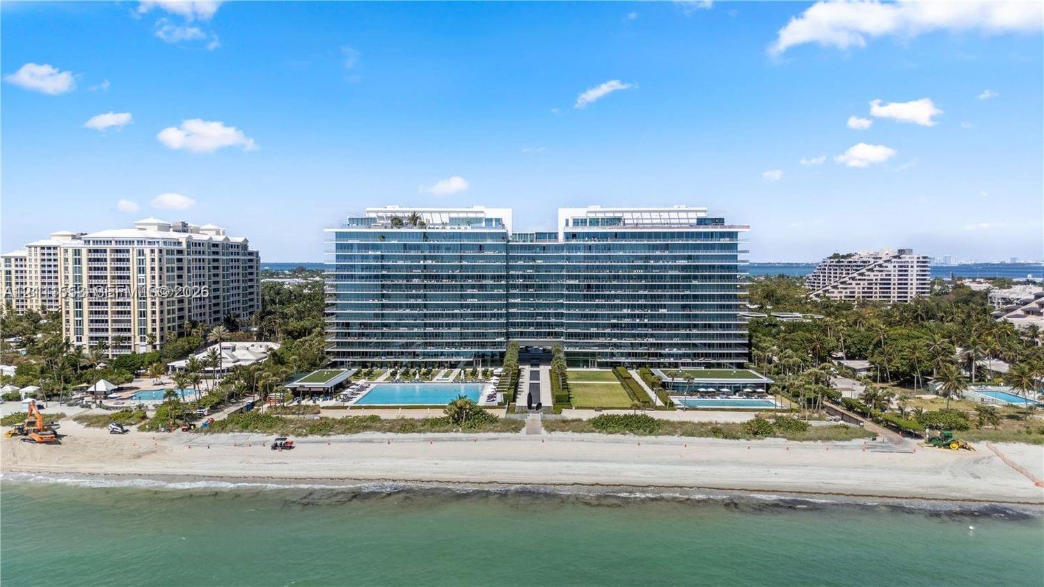 High-rise residential building featuring floor-to-ceiling glass paneling, expansive balconies, ground-level swimming pools, landscaped lawn areas, and direct beach access