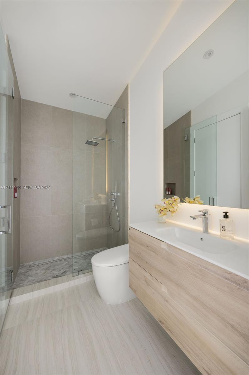 Floating wood-finish vanity with an integrated basin, frameless glass walk-in shower featuring a rainfall showerhead, neutral stone-finish tiling, and an expansive wall-mounted mirror with integrated backlighting