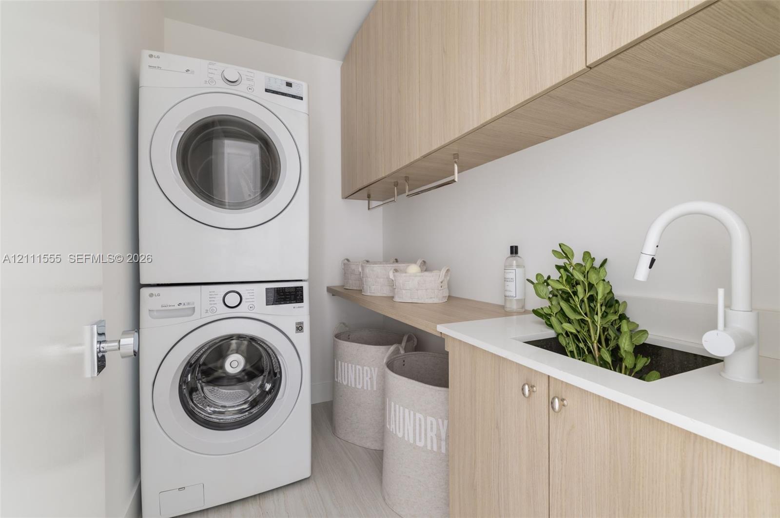Laundry space featuring stacked front-load laundry appliances, light wood-finish cabinetry, an under-mount sink, white solid-surface countertop, and wall-mounted white faucet
