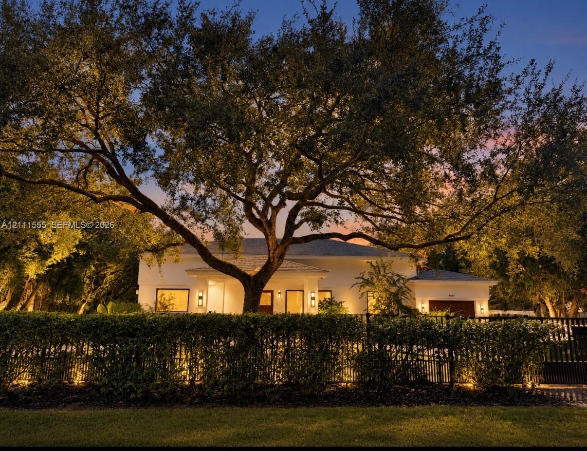 Single-story residence featuring light-colored exterior finish, mature specimen trees, perimeter metal fencing, and integrated outdoor lighting Single-story residence featuring light-colored exterior finish, mature specimen trees, perimeter metal fencing, and integrated outdoor lighting