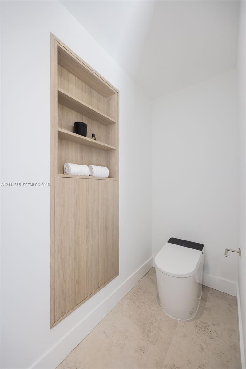 Built-in recessed wood-finish cabinetry featuring open shelving and lower storage cabinets, paired with a modern one-piece toilet and neutral stone-finish flooring