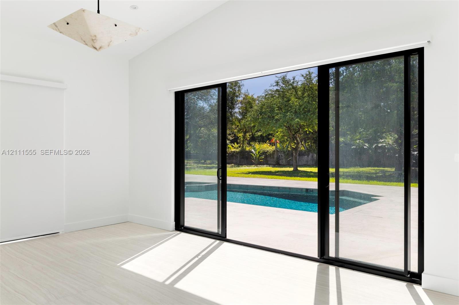 Vaulted ceiling, geometric pendant lighting, light-toned flooring, and expansive black-framed sliding glass doors opening to an outdoor swimming pool and patio