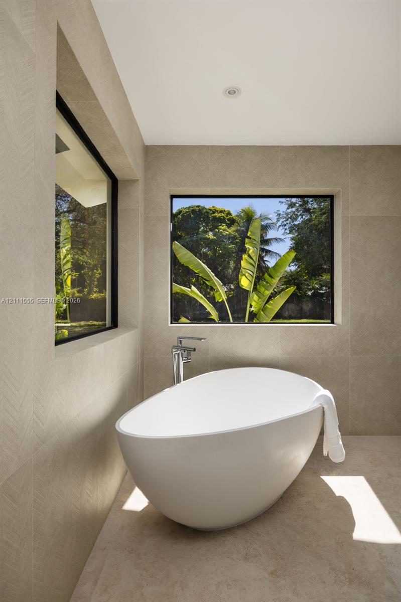 Freestanding soaking tub featuring a chrome floor-mounted faucet, set against large-format neutral tile walls with textured herringbone detailing and recessed windows