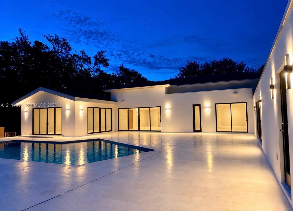 Contemporary exterior featuring smooth stucco finish, expansive stone patio, integrated wall-mounted lighting, multiple sliding glass door sets, and a rectangular swimming pool