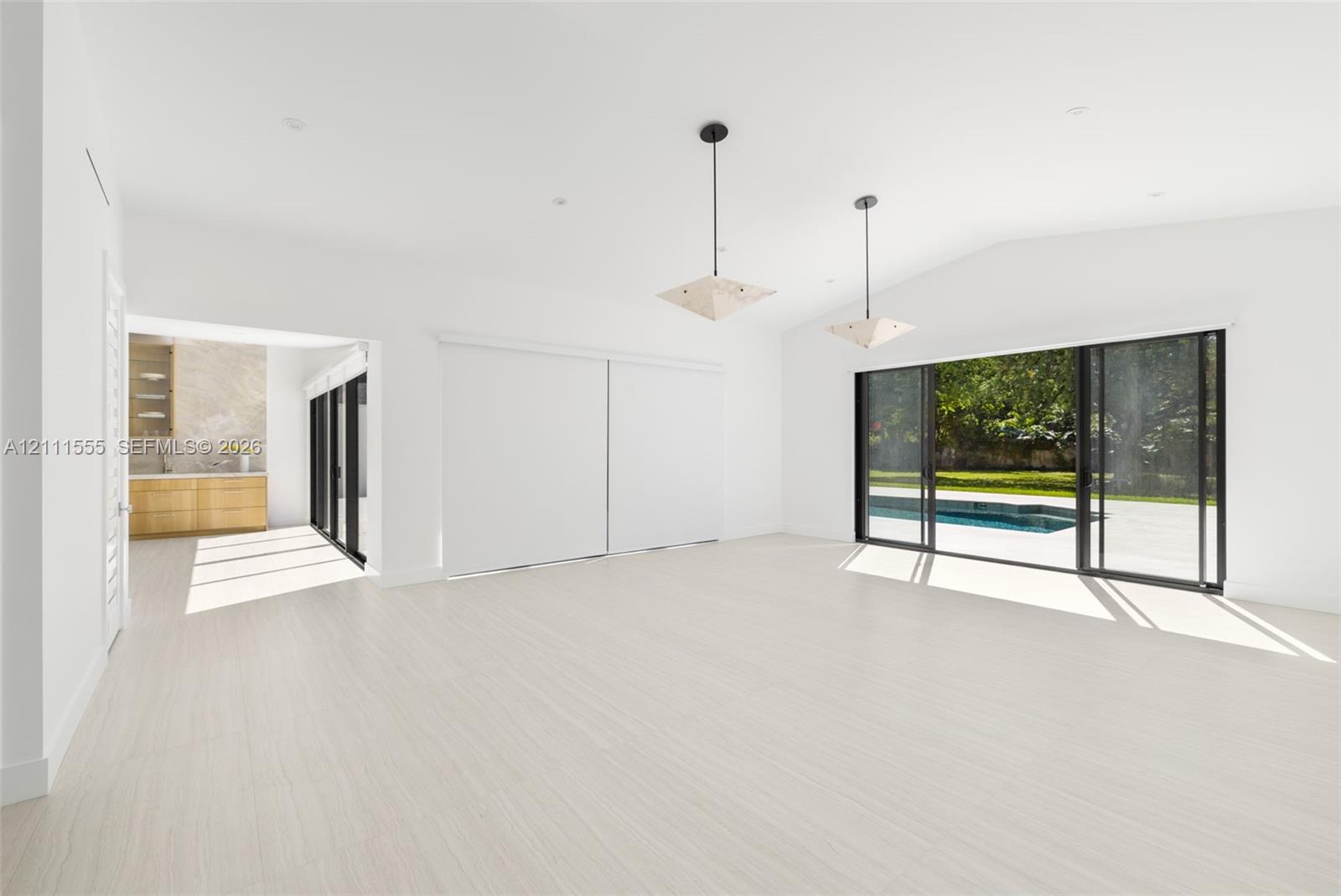 Open living area featuring vaulted ceilings, wood-finish flooring, dual geometric pendant lighting, and multi-panel sliding glass doors with exterior pool access
