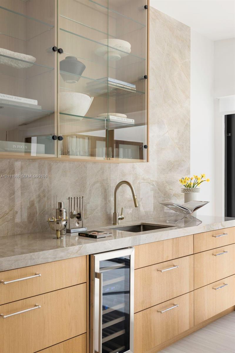 Wet bar featuring light-toned wood cabinetry, stone slab backsplash and countertop, under-counter wine refrigerator, and upper glass-front cabinetry with interior shelving Wet bar featuring light-toned wood cabinetry, stone slab backsplash and countertop, under-counter wine refrigerator, and upper glass-front cabinetry with interior shelving