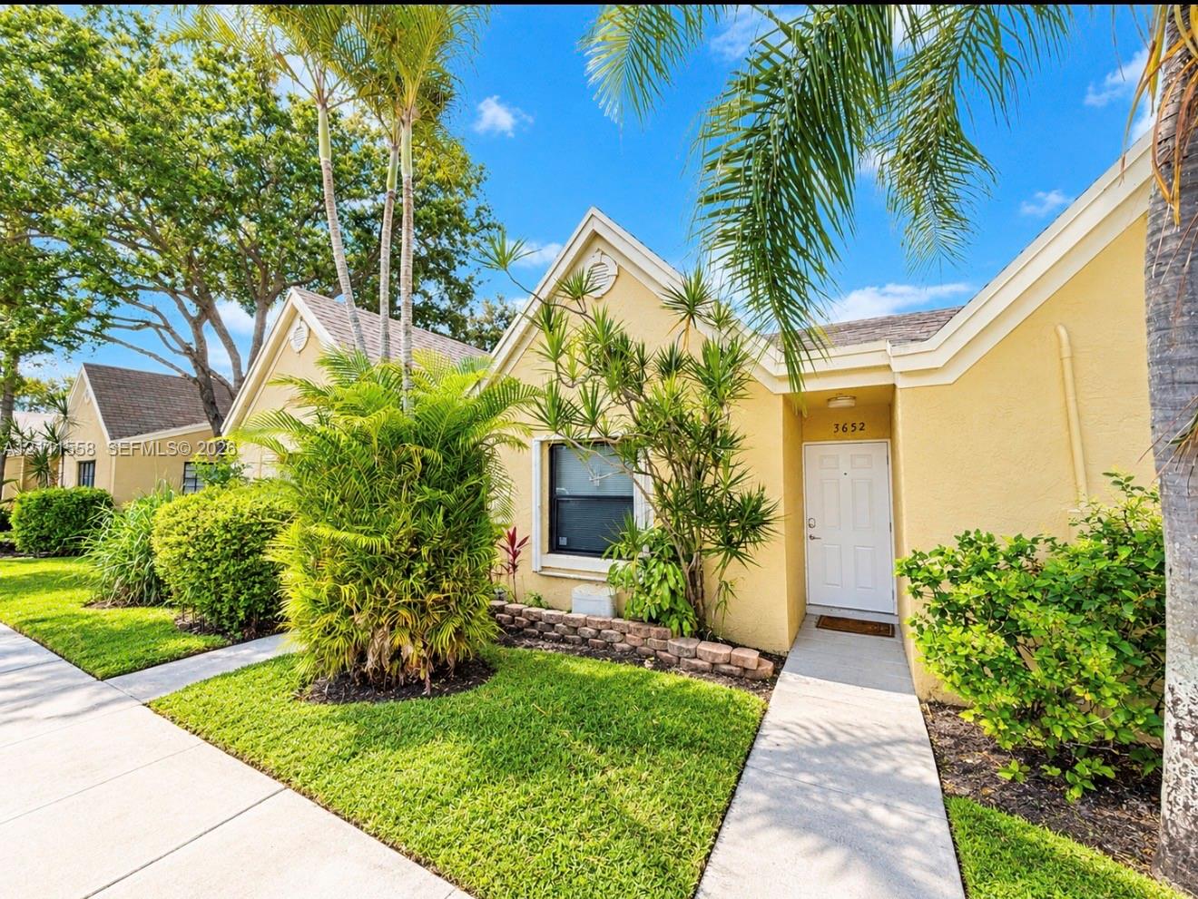 Stucco exterior with multi-gabled roofline, concrete walkway, and landscaped garden beds featuring mature palm trees and shrubs