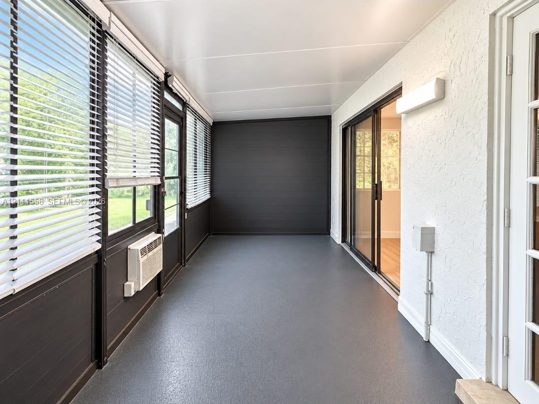 Enclosed sunroom featuring expansive windows with horizontal blinds, textured white wall finishes, a solid flooring surface, a sliding glass door, and a dedicated wall-mounted climate control unit