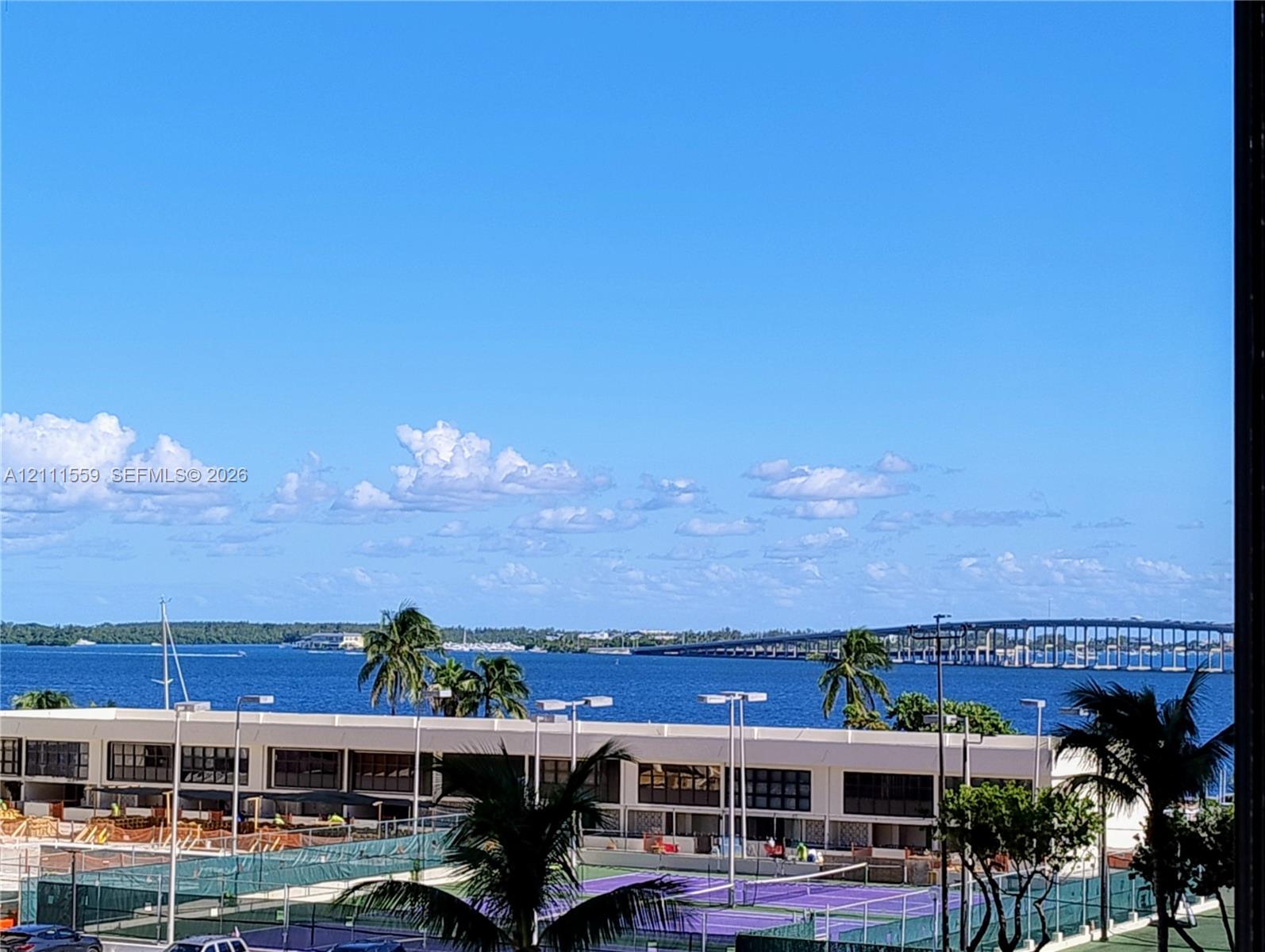 Open water backdrop featuring a bridge, palm trees, and outdoor tennis courts