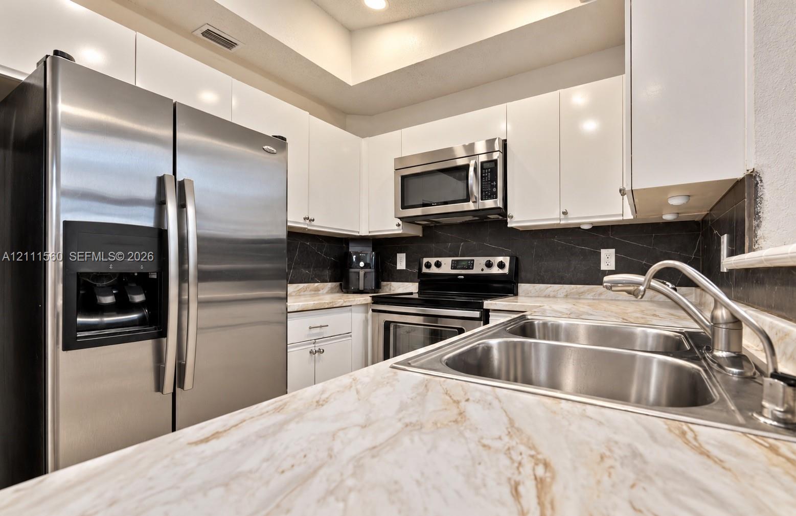 Kitchen featuring white gloss cabinetry, stone-patterned countertops, stainless steel appliances, and a dark stone-finish backsplash with an integrated dual-basin stainless steel sink