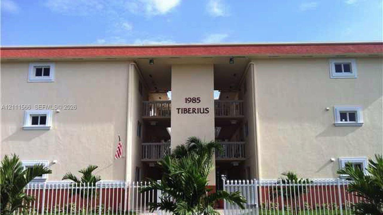 Exterior facade featuring multi-story balconies, stucco finish, a decorative roofline, and perimeter white metal fencing