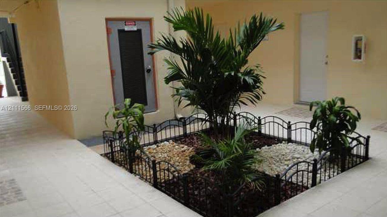 Landscaped courtyard featuring light-toned tile flooring, a central garden bed with ornamental shrubs, decorative gravel ground cover, and a black wrought-iron perimeter fence Landscaped courtyard featuring light-toned tile flooring, a central garden bed with ornamental shrubs, decorative gravel ground cover, and a black wrought-iron perimeter fence