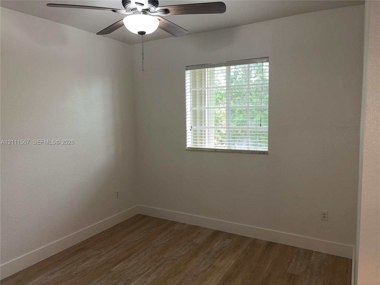 Interior room featuring wood-finish flooring, a multi-blade ceiling fan with integrated light fixture, and a single-pane window with horizontal blinds