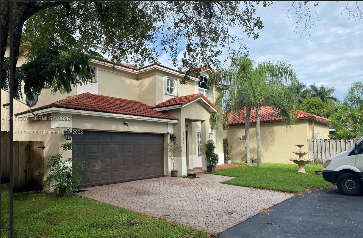Stucco exterior residence featuring a multi-gabled tile roof, paver driveway, attached two-car garage, and tiered garden fountain Stucco exterior residence featuring a multi-gabled tile roof, paver driveway, attached two-car garage, and tiered garden fountain