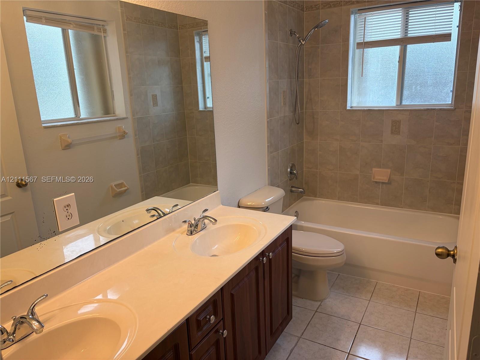 Double-sink vanity with a solid-surface countertop, dark wood-finish cabinetry, ceramic tile flooring, and a shower-tub combination featuring full-height wall tiling