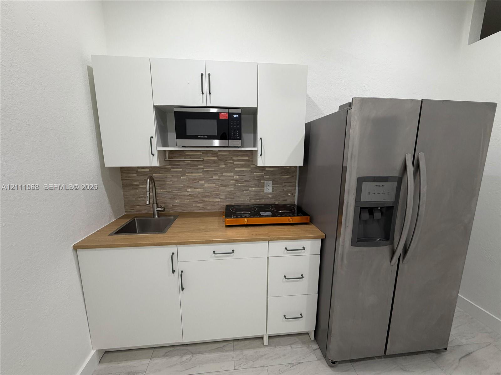 Kitchenette featuring white cabinetry, light wood-finish countertop, stainless steel sink with high-arc faucet, tile backsplash, and full-size stainless steel refrigerator