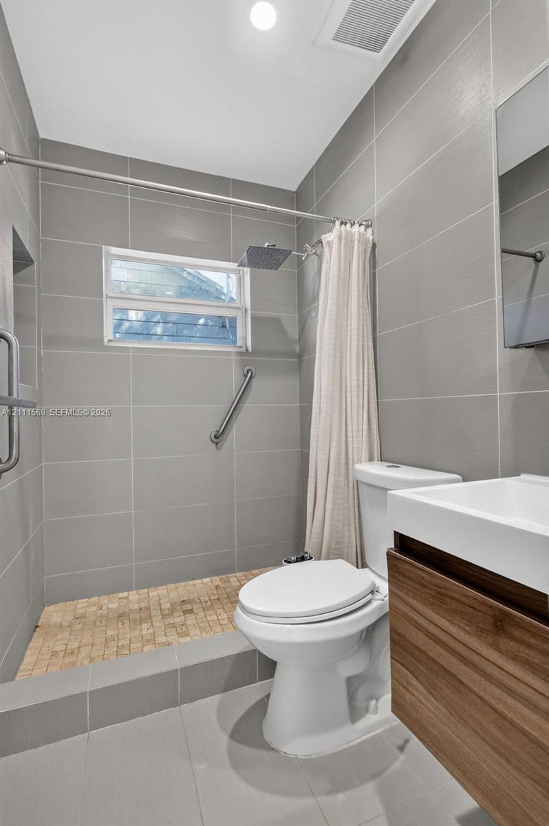Bathroom featuring floor-to-ceiling grey tile walls, a walk-in shower with mosaic tile flooring, a wood-finish floating vanity, and a rectangular vessel sink