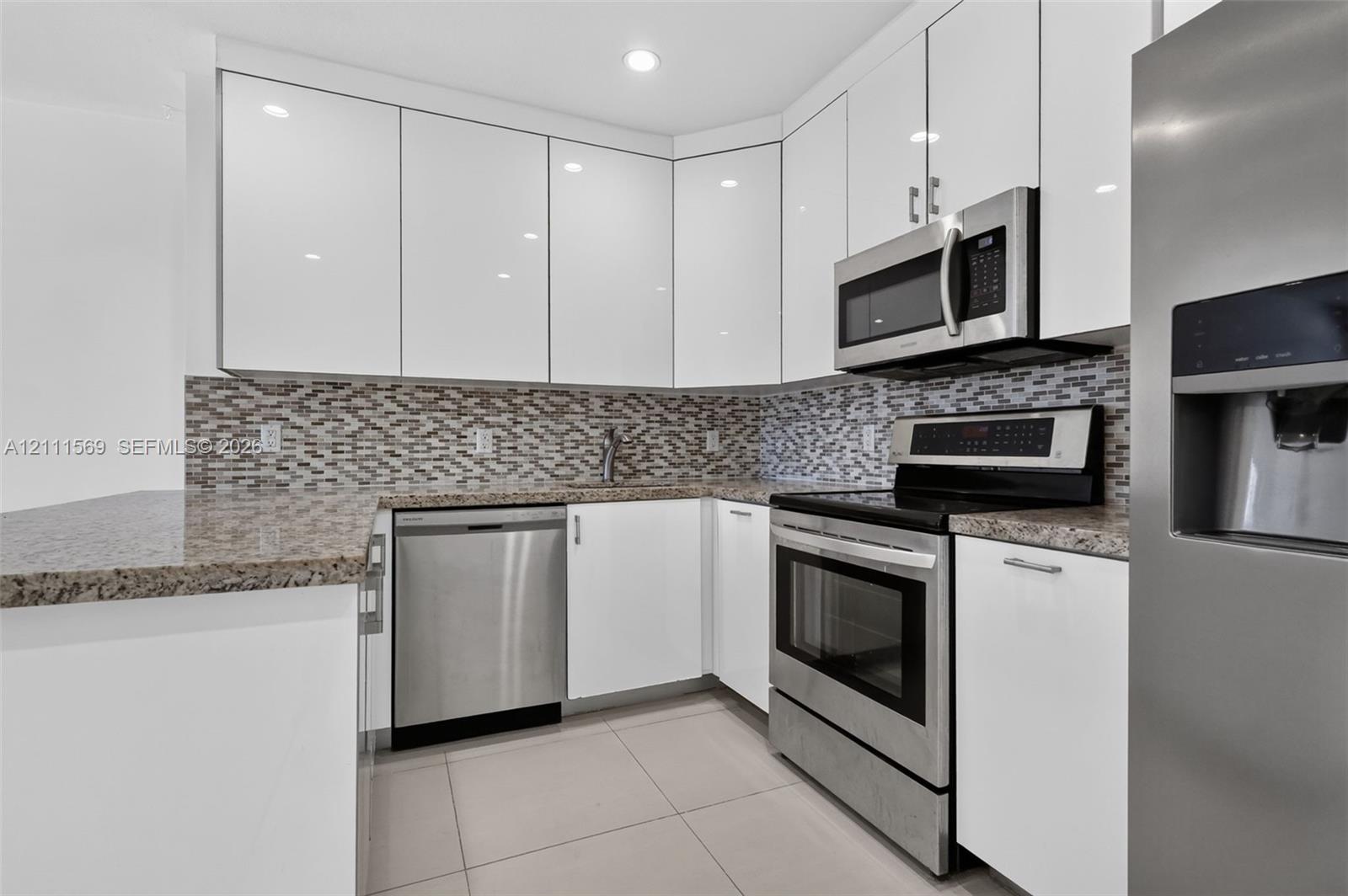 Kitchen featuring high-gloss white cabinetry, stone-finish countertops, stainless steel appliances, and a glass-tile mosaic backsplash Kitchen featuring high-gloss white cabinetry, stone-finish countertops, stainless steel appliances, and a glass-tile mosaic backsplash