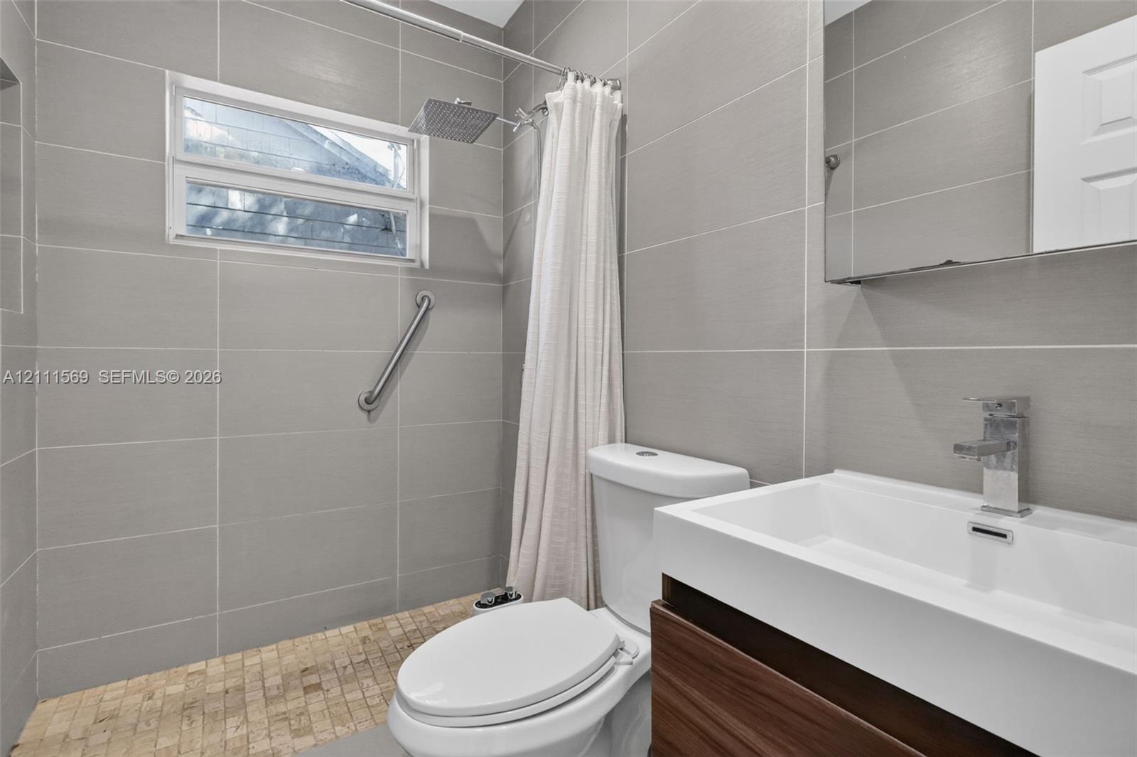 Bathroom featuring large-format gray wall tile, wood-finish vanity with an integrated rectangular sink, wall-mounted rain shower head, and textured stone floor tile