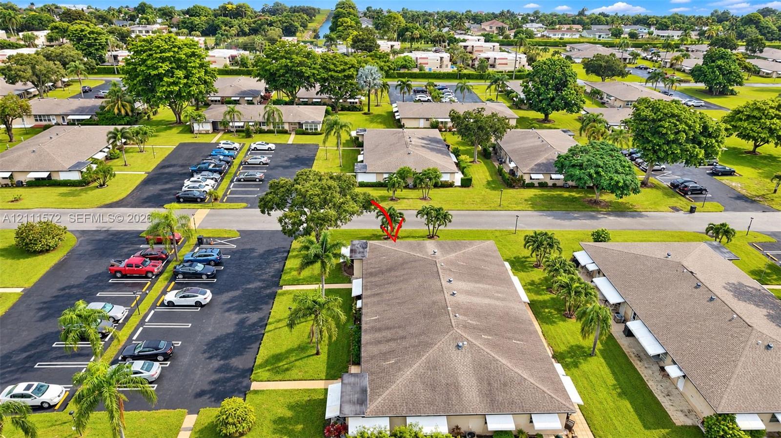 Aerial perspective of a well-maintained residential complex featuring low-rise structures with shingle roofing, paved surface parking, and extensive green space with mature palm trees