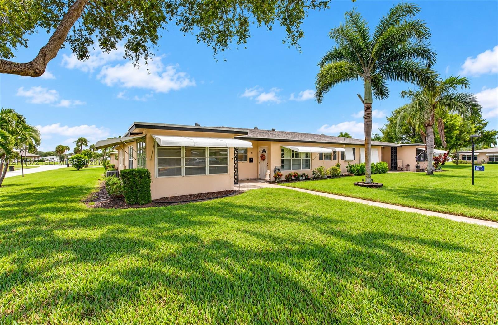 Single-story residence featuring light-toned exterior finish, retractable window awnings, and palm-accented landscaping with expansive green space