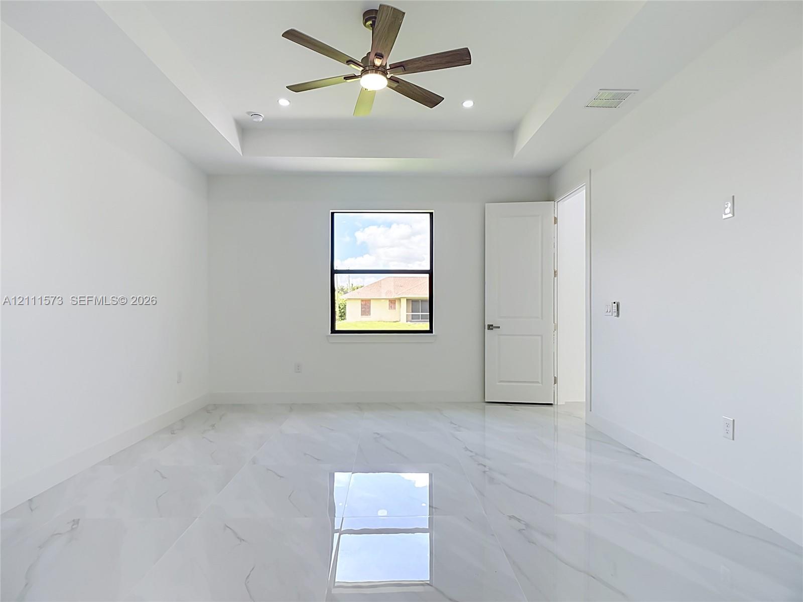 Tray ceiling, polished tile flooring with veining, multi-blade ceiling fan with integrated lighting, and dark-framed window