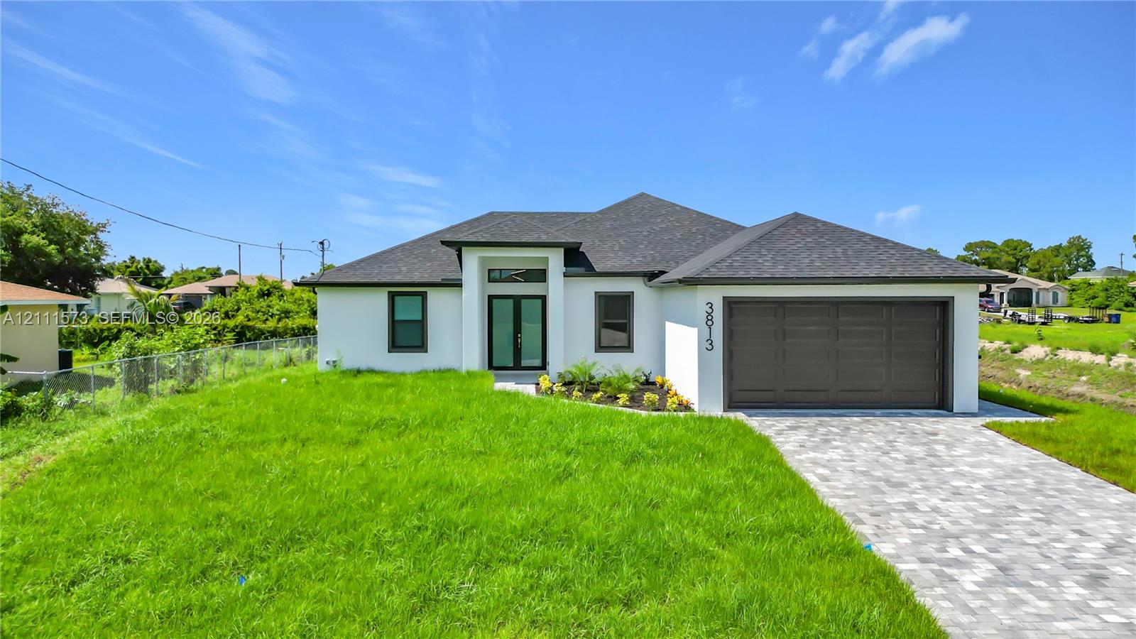 Single-story exterior featuring white stucco siding, a grey shingle roof, double-entry doors with transom window, and a paver driveway leading to an attached two-car garage