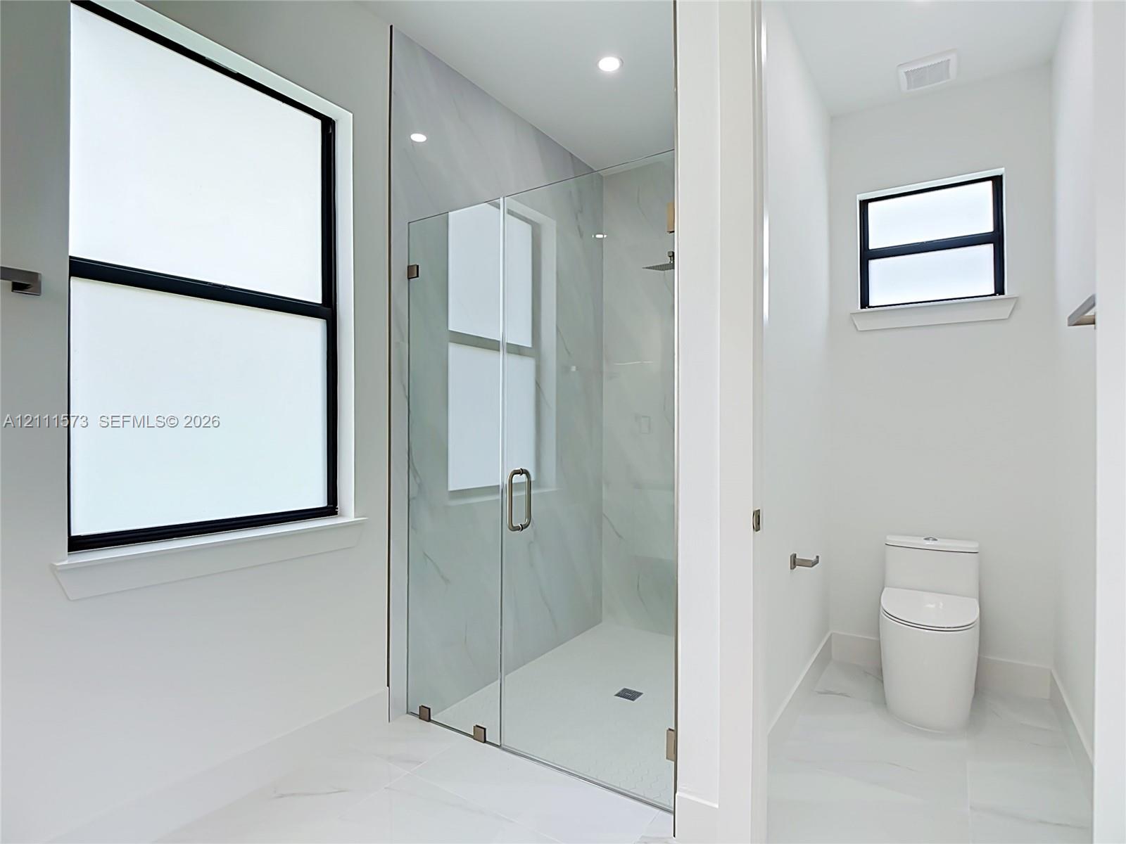 Walk-in shower featuring glass enclosure, stone-finish wall tile, and recessed lighting, accompanied by a low-profile toilet and black-framed windows