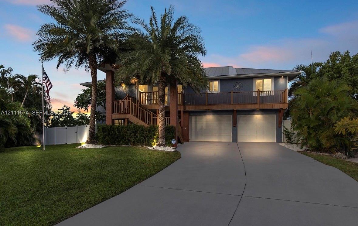 Two-story residence featuring a metal roof, attached two-car garage, elevated wooden staircase, wrap-around balcony with wooden railings, and mature palm landscaping