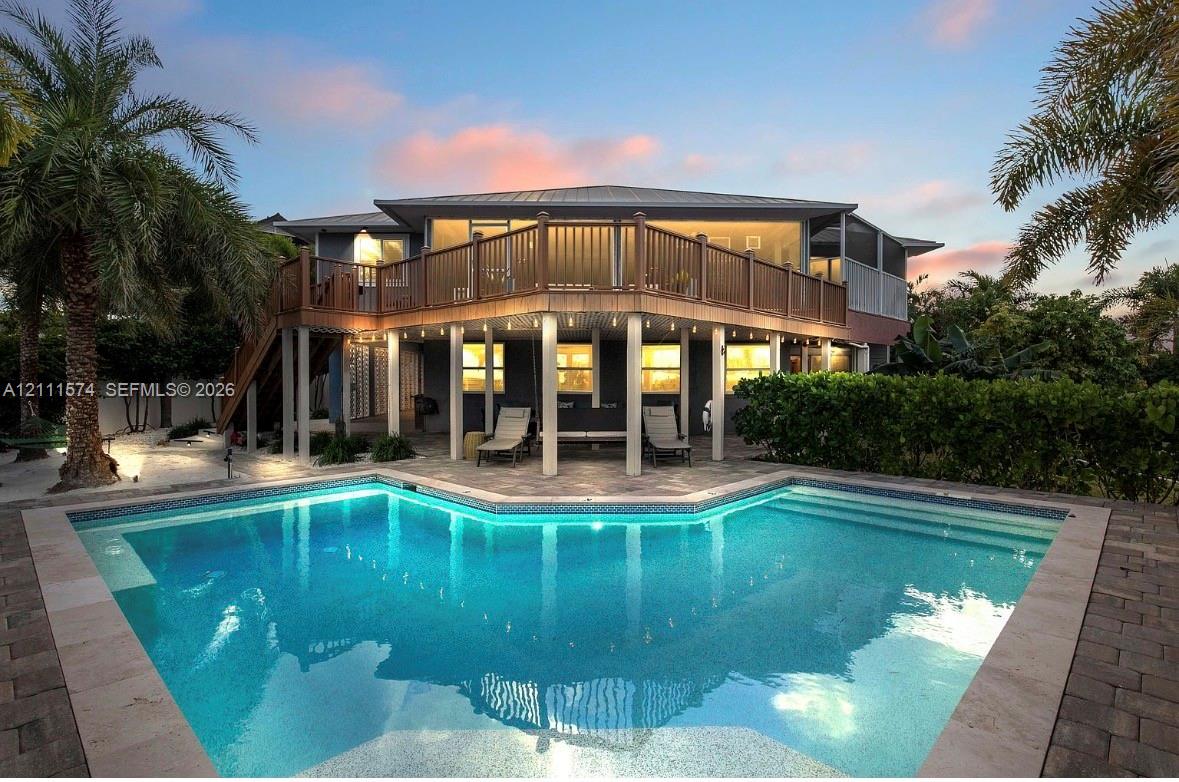 Multi-level elevated home featuring a wrap-around wooden deck, a ground-level covered patio area with support columns, and an inground swimming pool with stone-style paver decking