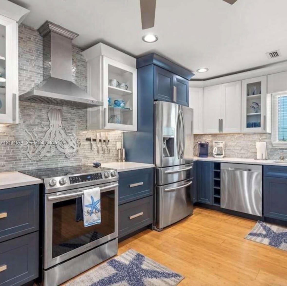 Kitchen featuring two-tone cabinetry, stainless steel appliances, a glass-tile backsplash with decorative accent, solid surface countertops, and wood-finish flooring