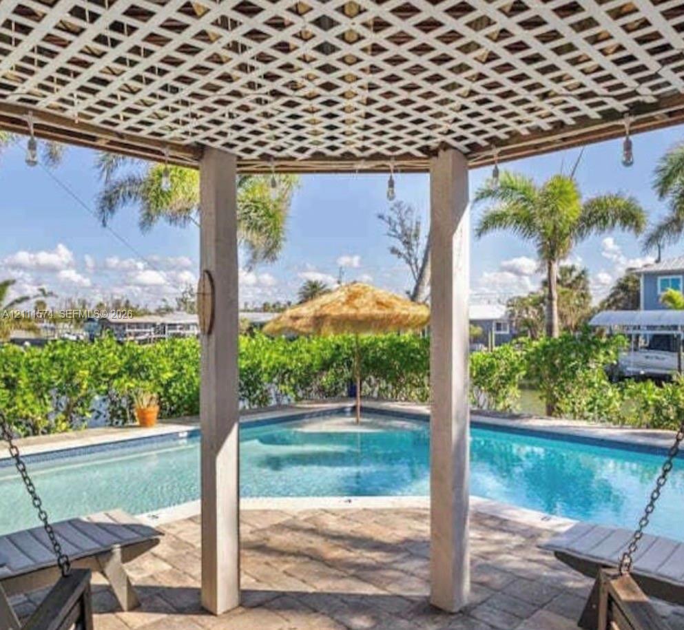 Covered gazebo featuring a lattice roof, stone paver flooring, and suspended bench seating, positioned over a swimming pool with water access