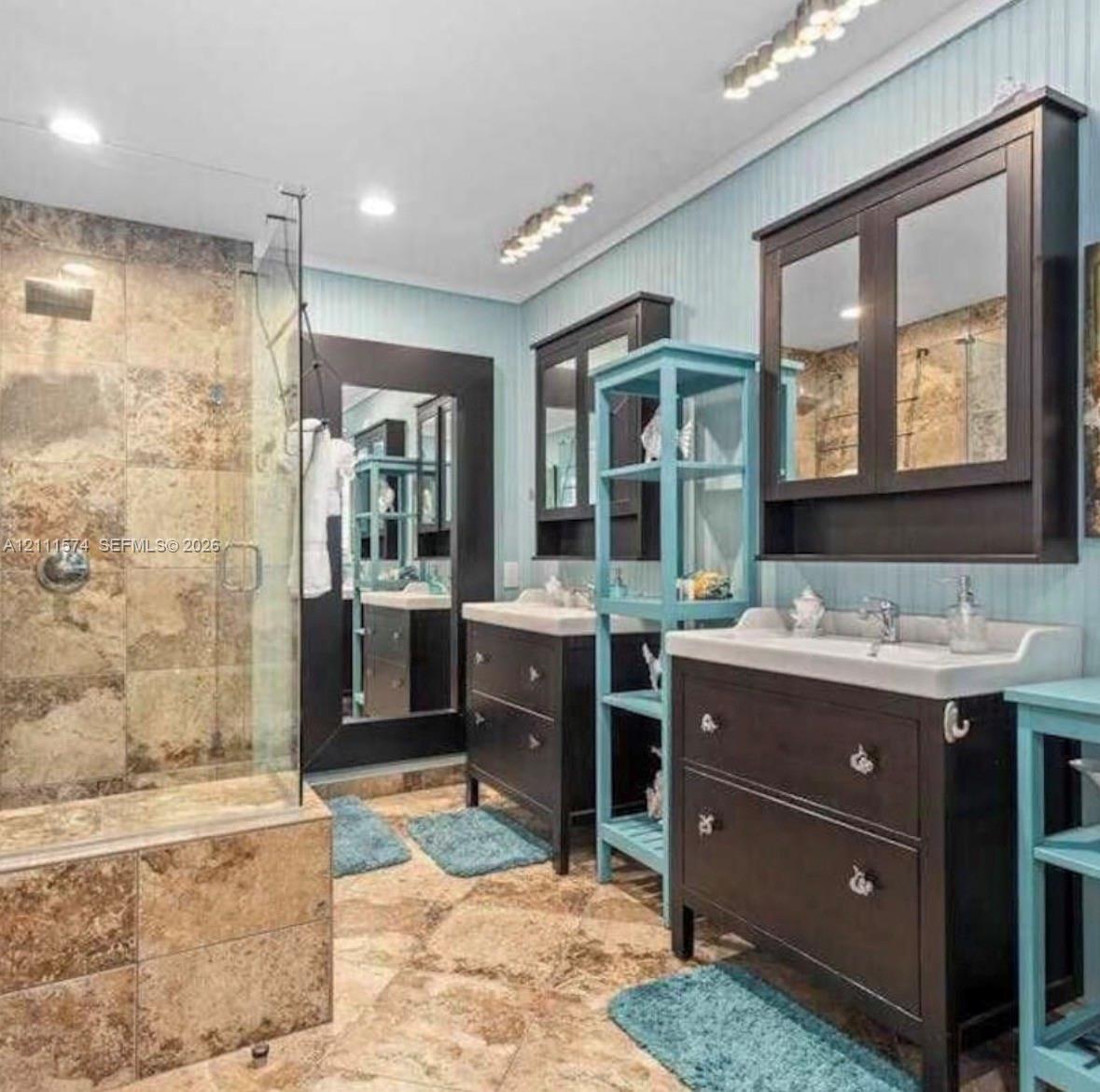 Bathroom featuring a walk-in shower with stone tile surround, dual dark-finish vanities with integrated cabinetry, beadboard wall paneling, and stone tile flooring