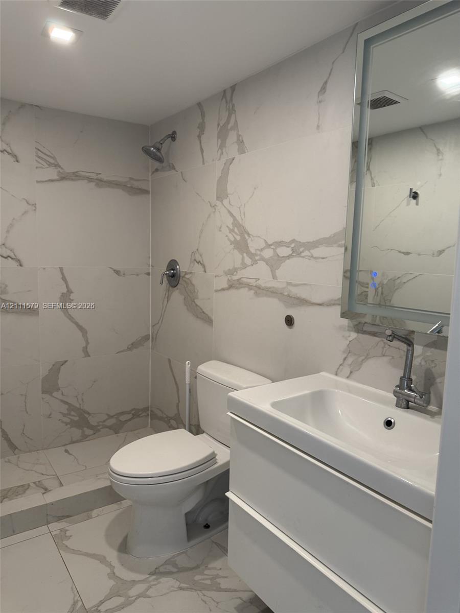 Full-height wall tiling with a vein-pattern finish, modern white floating vanity, integrated rectangular sink, wall-mounted backlit mirror, and recessed overhead lighting