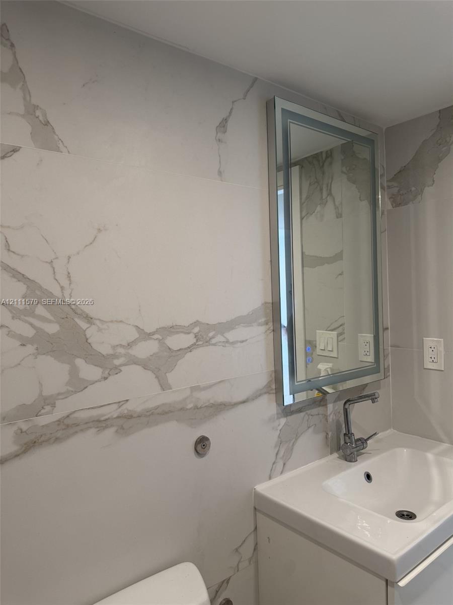 Large-format white stone-finish wall tile with grey veining, integrated mirror with illuminated border, and pedestal sink featuring a chrome faucet