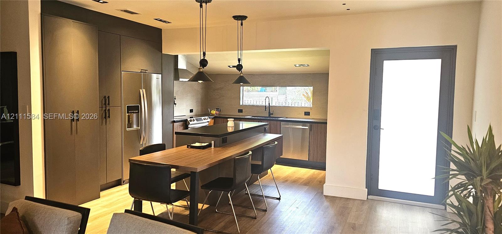 Modern kitchen featuring dark matte cabinetry, stainless steel appliances, a central island with an integrated wood-finish breakfast bar, and recessed lighting paired with industrial-style pendant lights