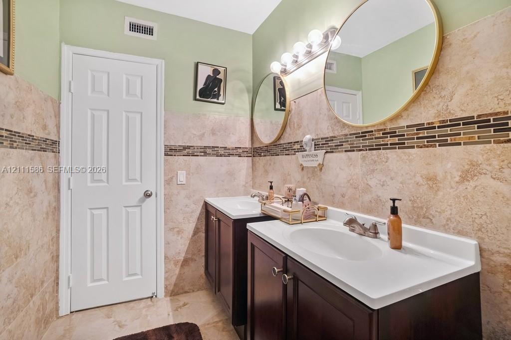 Dual vanity with dark wood-finish cabinetry, integrated white basins, stone-tiled walls with decorative mosaic accent inlay, and circular mirrors with metallic frames