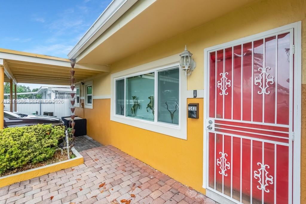 Exterior entryway featuring a paver stone walkway, attached carport, decorative metal security door, wall-mounted exterior light fixture, and chain-style rain gutter