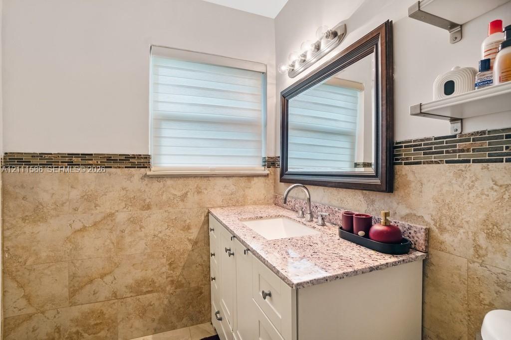 Bathroom vanity with stone countertop, undermount sink, multi-drawer white cabinetry, framed wall mirror, and decorative tile wainscoting