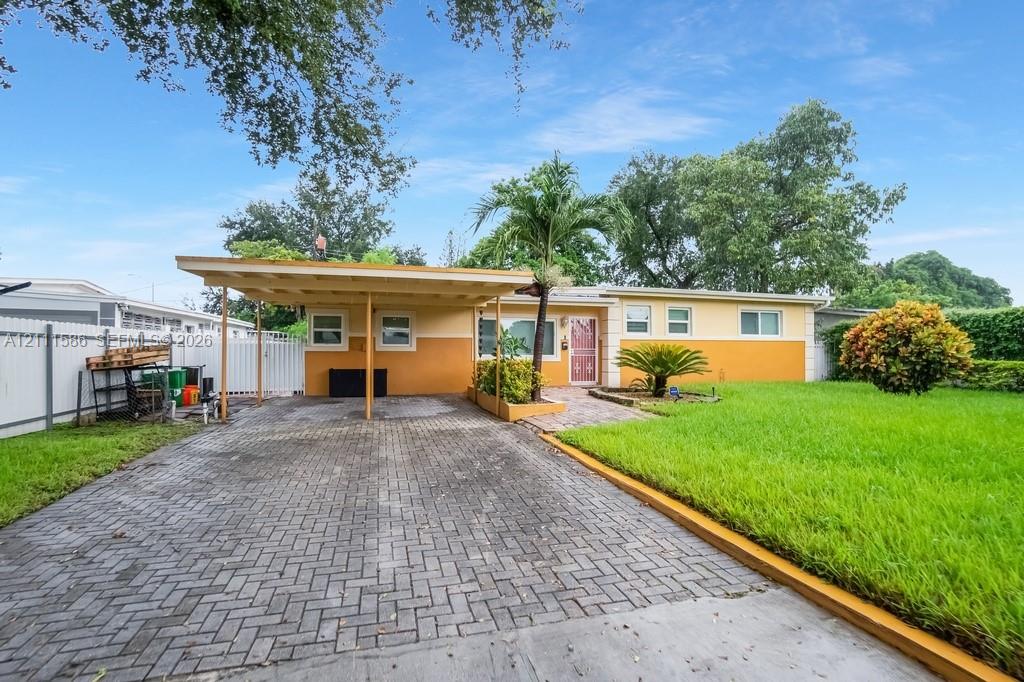 Single-story exterior featuring an attached carport, herringbone-patterned paved driveway, front lawn, and vinyl privacy fencing