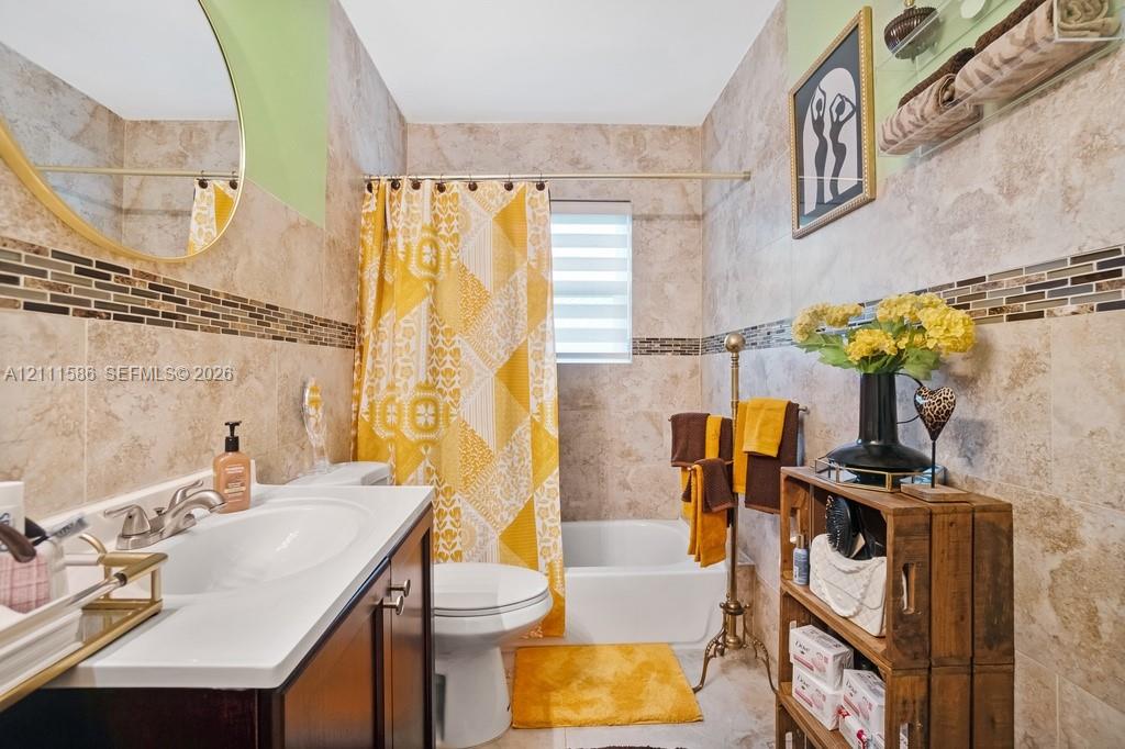 Full-wall tile surround featuring decorative mosaic accent tiling, vanity with an integrated white sink, circular mirror with metallic frame, and tiered horizontal blind
