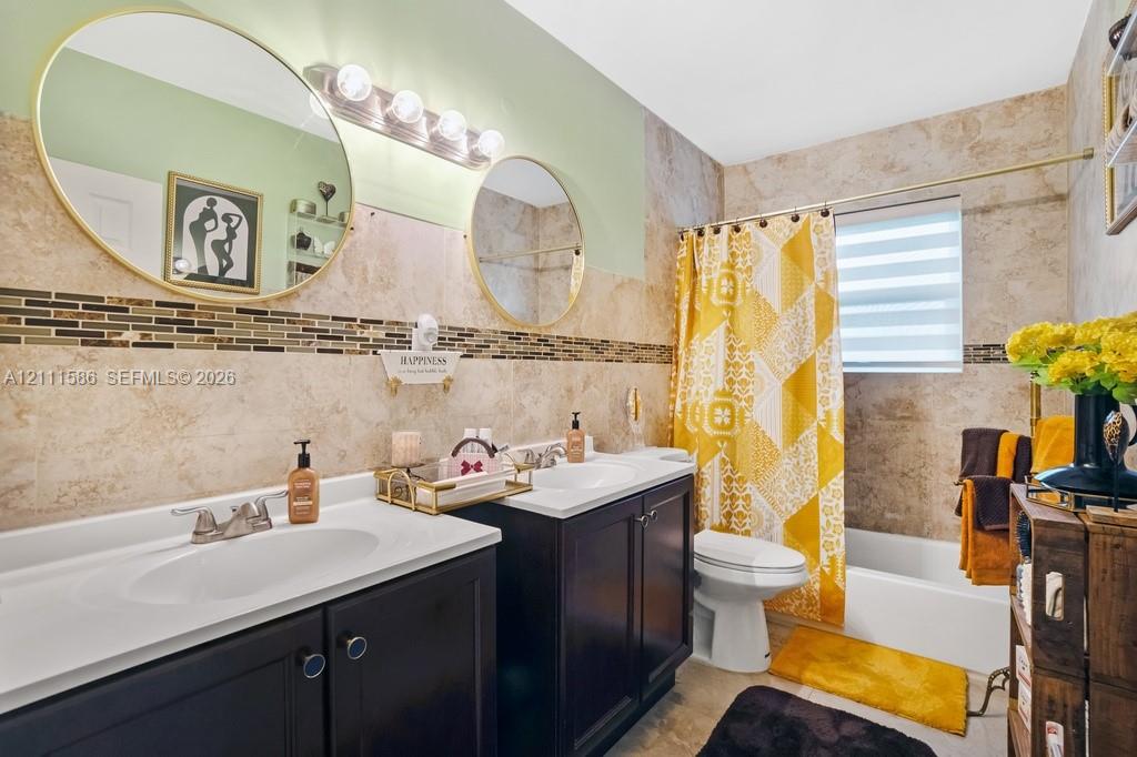 Dual dark-finish vanities with integrated sinks, large round wall-mounted mirrors, and tiled shower surround with decorative mosaic accent inlay