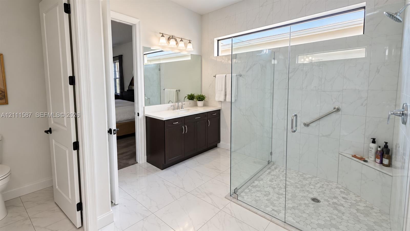 Walk-in shower with stone-finish tile surround, frameless glass enclosure, and built-in bench seating, complemented by a dark wood vanity with white countertop and large vanity mirror