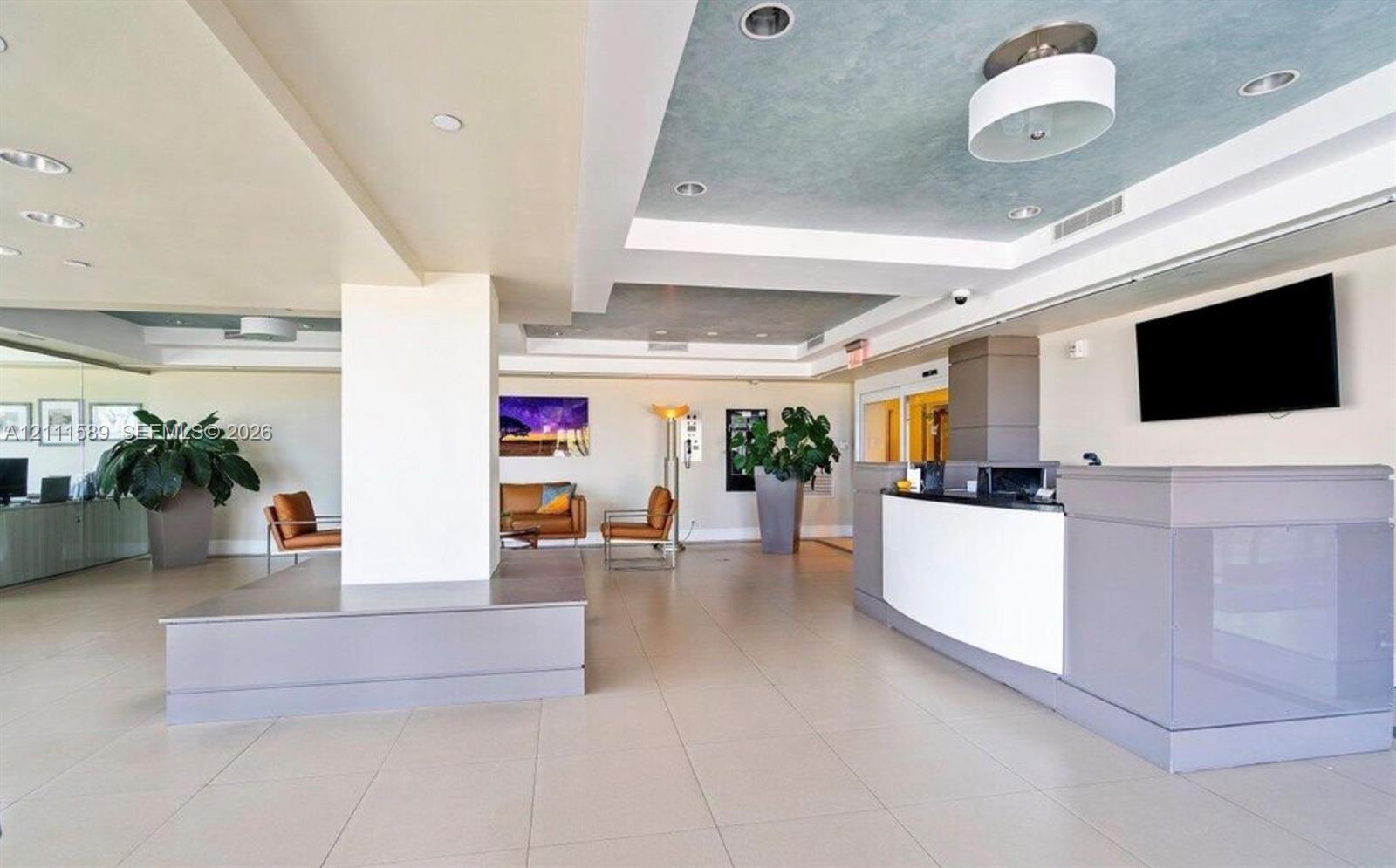 Reception area featuring large-format tile flooring, a tiered tray ceiling with recessed lighting, a structured built-in service counter, and an integrated central pillar with a stepped platform base Reception area featuring large-format tile flooring, a tiered tray ceiling with recessed lighting, a structured built-in service counter, and an integrated central pillar with a stepped platform base