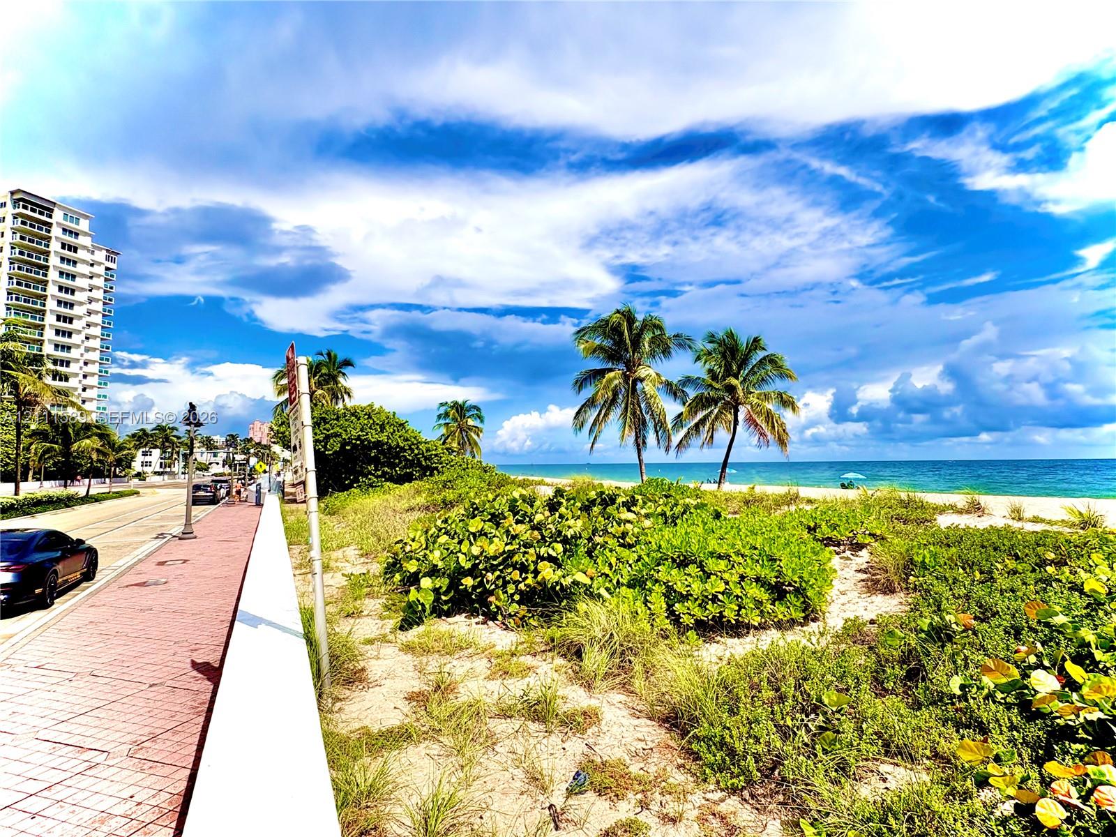 Coastal property featuring a brick-paved sidewalk, decorative street lamps, mature palm trees, native beach vegetation, and direct oceanfront access