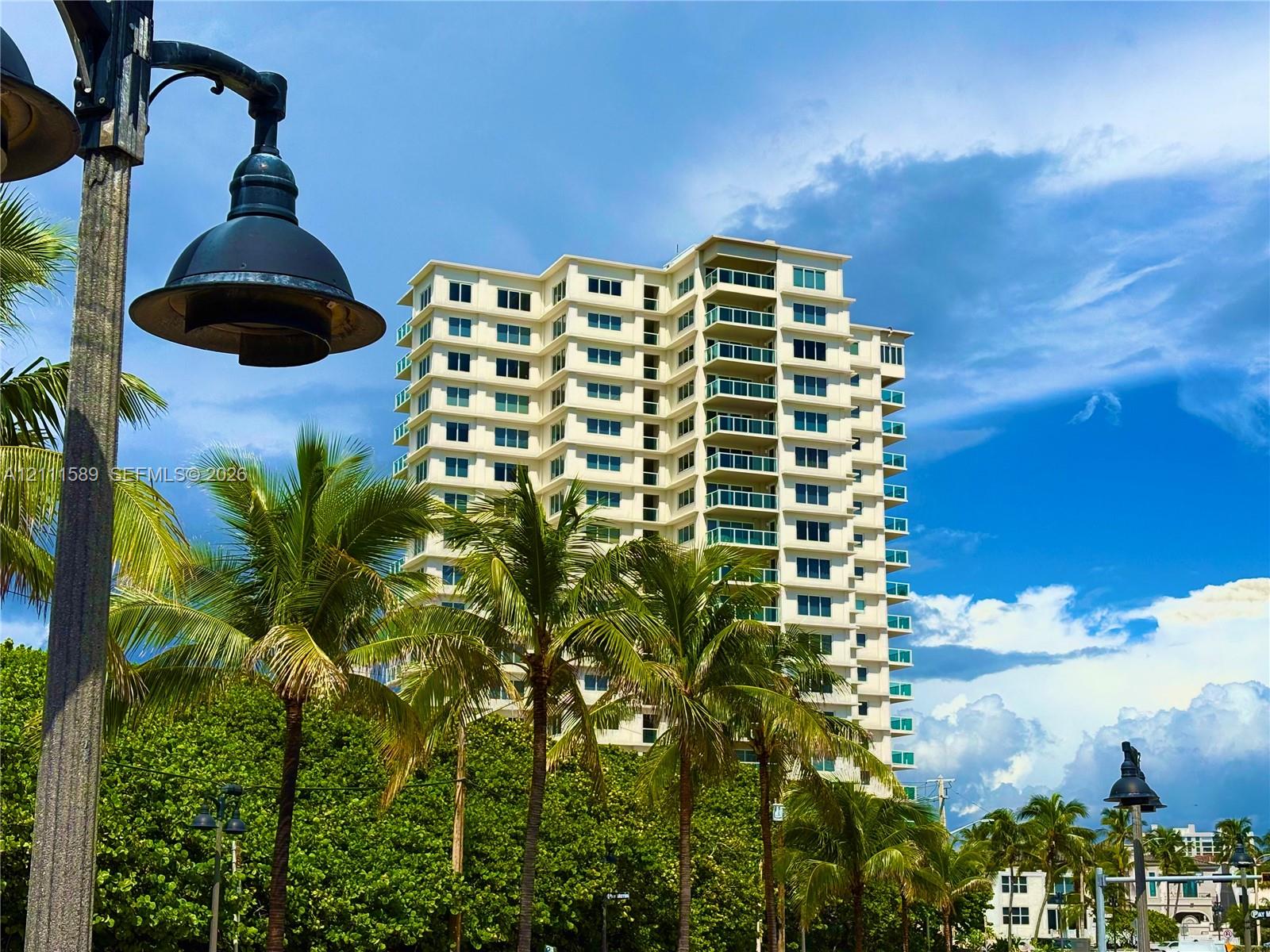 High-rise residential building featuring multi-level private balconies with glass railings, light-toned exterior facade, and mature landscaping