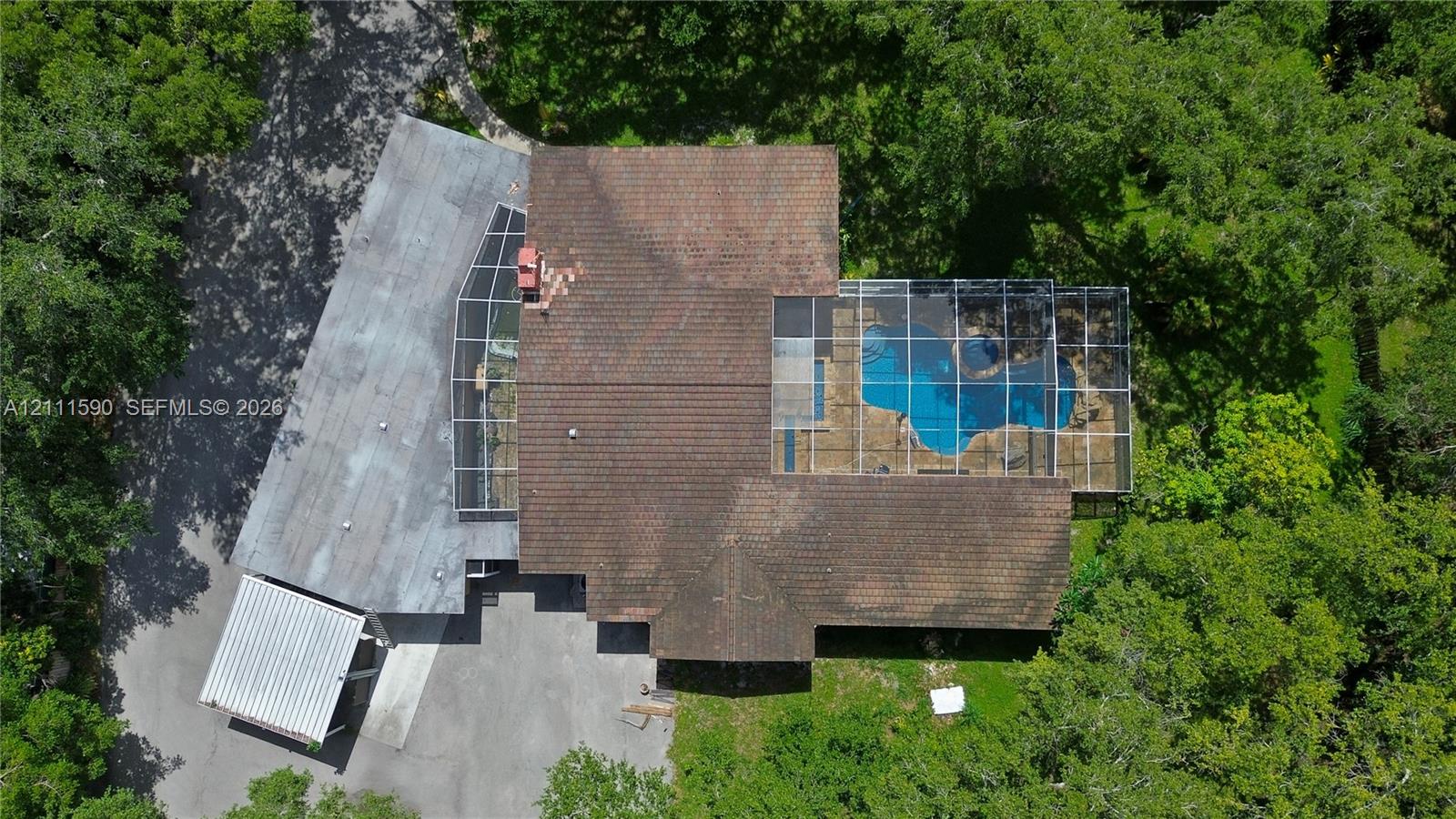 Aerial perspective featuring a multi-level roofline, screened-in swimming pool enclosure, detached covered parking structure, and mature trees on a wooded lot