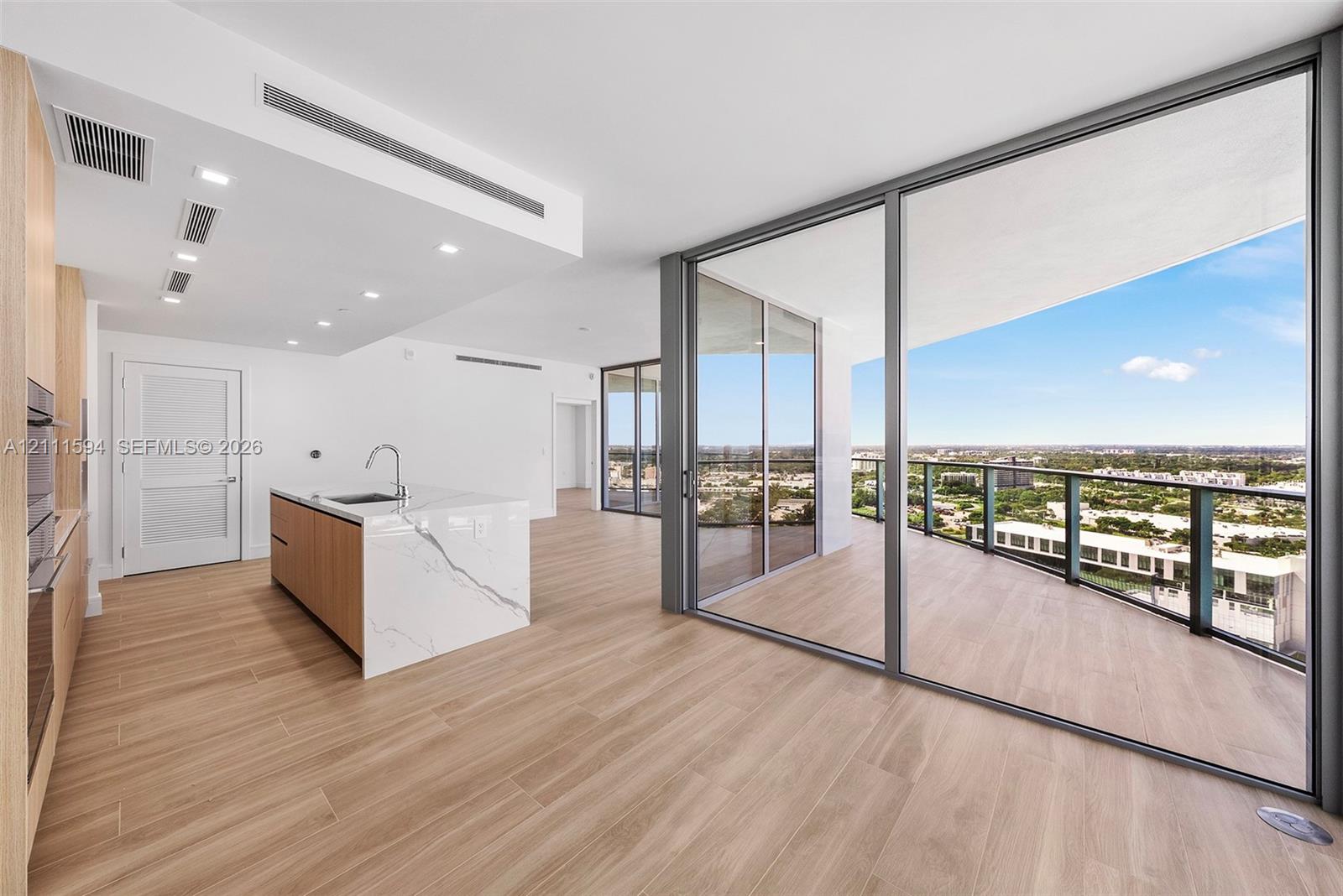 Kitchen island with a stone-finish waterfall edge and integrated sink, floor-to-ceiling glass sliding doors, expansive covered balcony, and wood-finish flooring throughout