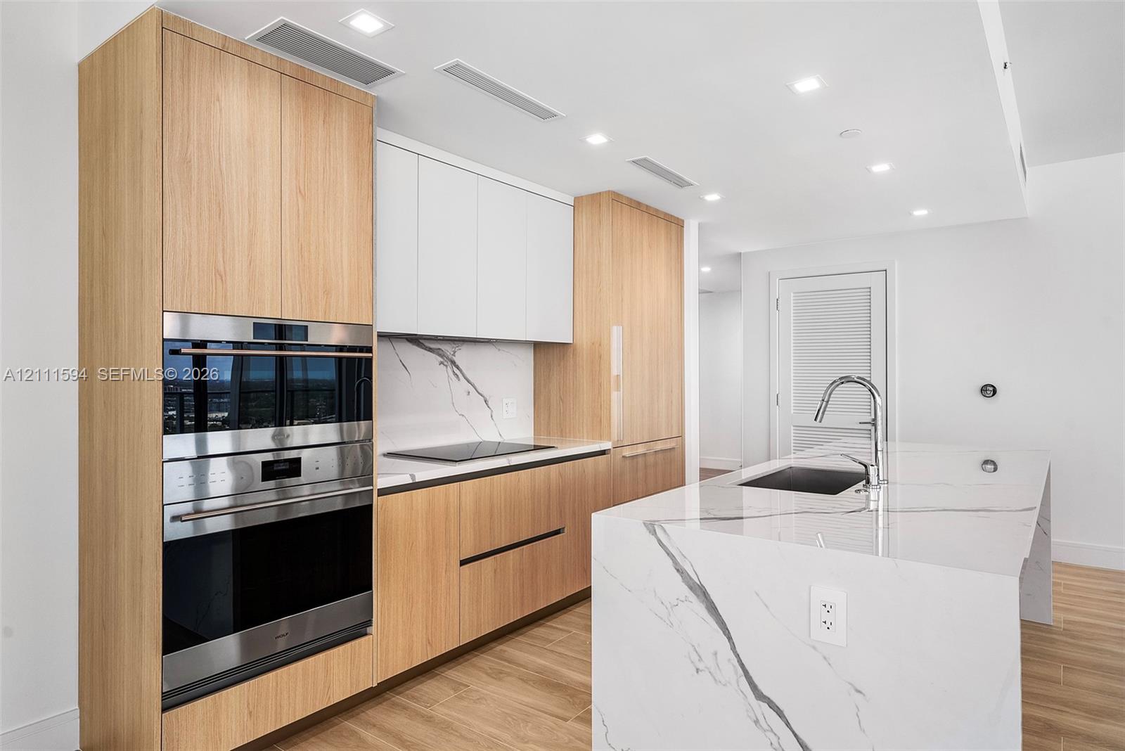 Kitchen featuring wood-finish cabinetry, white slab-style upper cabinets, stone-surfaced island with waterfall edge, integrated stainless steel oven and microwave, and recessed ceiling lighting